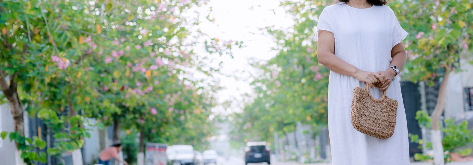 Femme en robe blanche avec sac en paille dans une rue fleurie — accessoires d'été Vogue Française